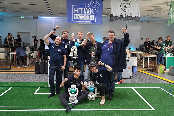 Das Team in Hamburg. (Foto: HTWK Robots) Menschen mit humanoiden Robotern auf kleinem Fußballfeld indoor