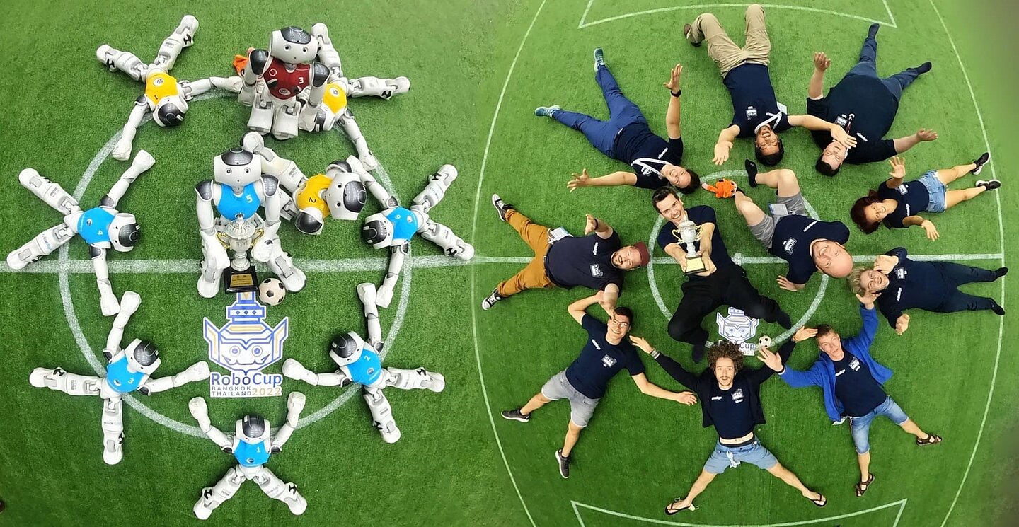 Menschen und Roboter liegen auf grünem Grund und halten Pokal hoch