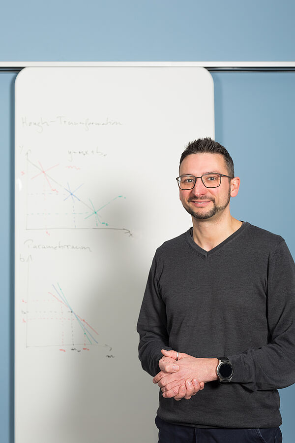 Dr. Mirco Fuchs steht vor einem Whiteboard, auf dem Skizzen für Algorithmen zu sehen sind