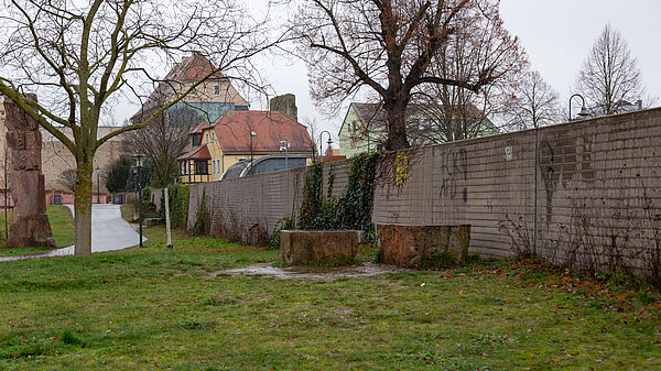 Eine Betonmauer, die teilweise mit Efeu bewachsen und Graffitti beschmiert ist. Vor der Mauer befindet sich eine Wiese mit einem Fußweg, hinter der Mauer sind Häuser.