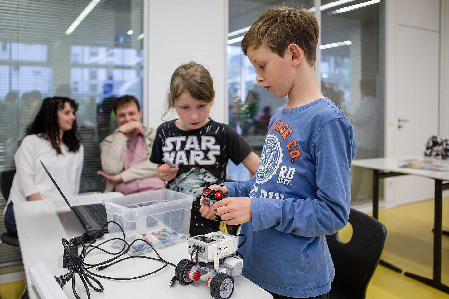 Wissenschaftsnacht 2018 Im Vordergrund zwei Kinder, die einen Mini-Roboter bauen, im Hintergrund zwei Erwachsene im Gespräch