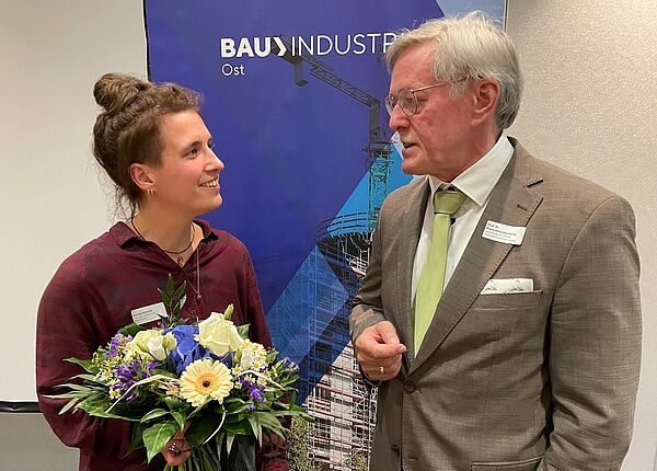 links steht eine Frau und rechts ein Mann, sie schauen sich an. Die Frau hält einen Blumenstrauß.