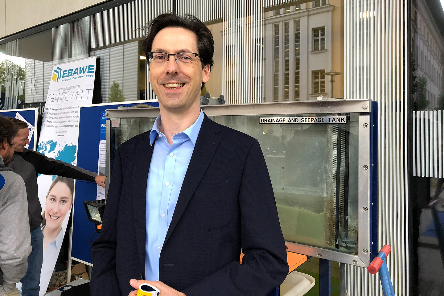 Wissenschaftsnacht 2018 Im Vordergrund eine in die Kamera lächelnde Person während im Hintergrund Menschen an einem Glasfenster-Element stehen