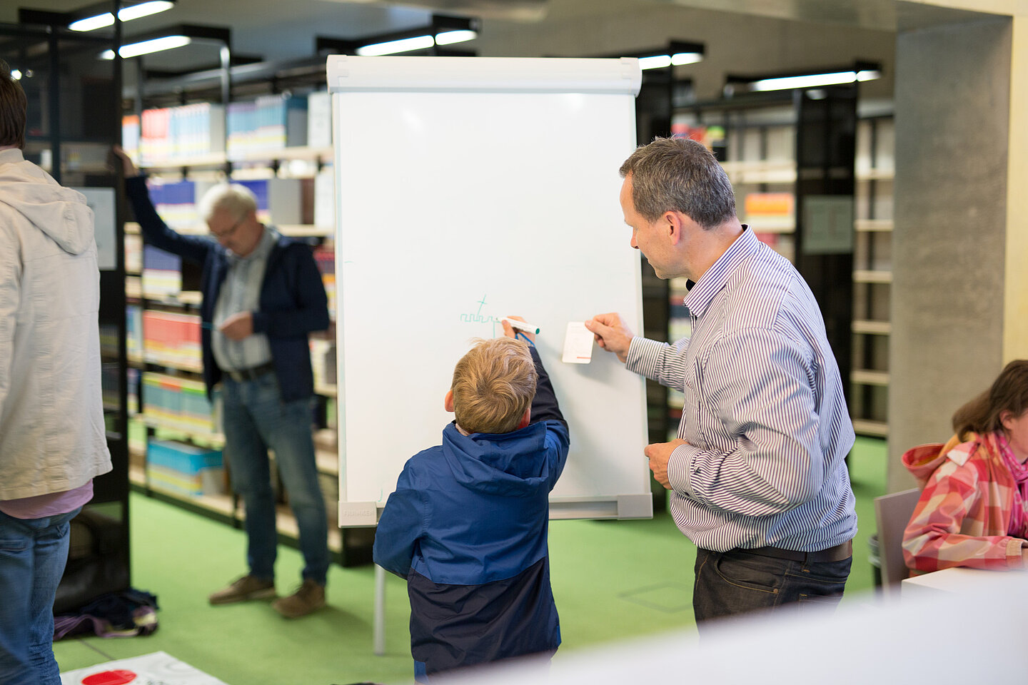 Wissenschaftsnacht 2018 Ein Kind und ein Erwachsender schreiben auf einem Flipchart, im Hintergrund Bücherregale und einzelne Menschen