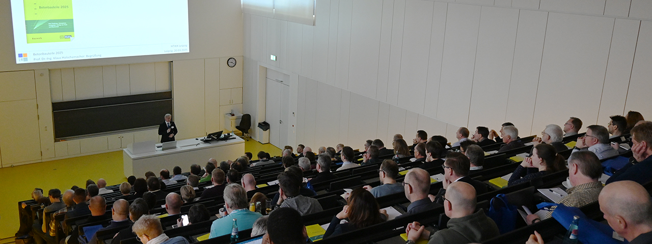 Ein großer Hörsaal mit ansteigenden Sitzreihen ist gut gefüllt mit Zuhörerinnen und Zuhörern. Vorne steht eine Person am Rednerpult und hält einen Vortrag. Über der Tafel ist eine Präsentationsfolie mit dem Titel „Betonbauteile 2025“ projiziert. Die Veranstaltung findet in einem modernen, hellen Vorlesungssaal statt.